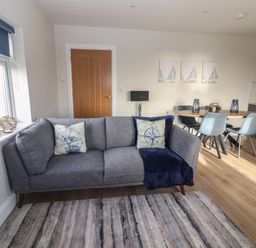 A living area with a gray sofa with pillows and blanket near a window and a dining table with chairs at The Boathouse in Carrigaholt County Clare