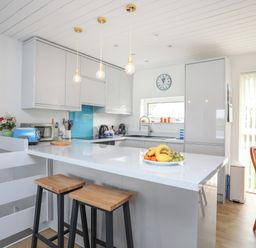 A kitchen with white cabinets a kitchen island with two stools and a fruit bowl at Waters Edge Ymyl Y Dwr in Y Felinheli