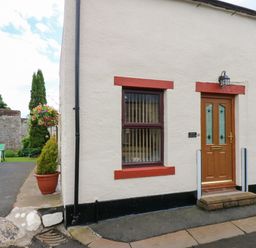 The exterior of a cottage with a wooden door and a window with red trim beside a small garden with green chairs at River Cottage in Eamont Bridge