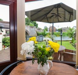 A wooden table with a vase of yellow and white flowers by windows overlooking a garden with chairs an umbrella and a river at River Cottage in Eamont Bridge