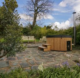 A garden patio with a wooden hot tub wooden steps and white metal benches surrounded by greenery at Eden Vale in Broughton-In-Furness