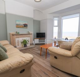 A living room with beige sofas a wooden coffee table a fireplace and a television near large windows overlooking the sea at 46 The Promenade in Withernsea