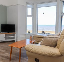 A living room with a TV on a stand a wooden coffee table and beige leather sofas next to large windows overlooking the sea at 46 The Promenade in Withernsea