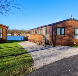 A gravel driveway leading to a brown wooden lodge near a lake with other lodges visible in the background at Gressingham 7 in Warton near Carnforth