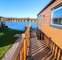 A wooden deck with railing next to a building by a lake with houses across the water at Gressingham 7 Warton near Carnforth
