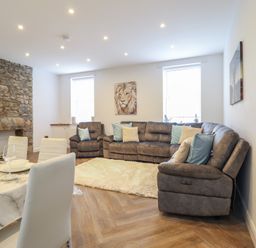 A living room with a stone fireplace a brown sectional sofa with pillows a white rug two windows and a dining table with white chairs at Gwydyr Bach 1 in Llanrwst