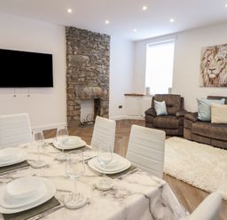 A dining area with a marble table and white chairs in an open living room with brown sofas and a stone fireplace at Gwydyr Bach 1 in Llanrwst