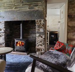 A living room with a wood burning stove a stone wall a round table and a sofa with blankets at Nyth Yr Eryr in Blaenau Ffestiniog