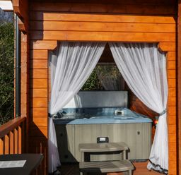 An outdoor hot tub on a wooden porch with sheer white curtains tied back at Little Lodge Balminnoch near Glenluce