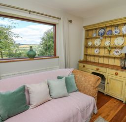 A living room with a sofa near a window and a yellow wooden cabinet with plates at Oak Apple Cottage in Presteigne