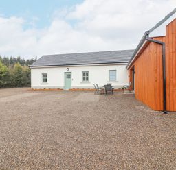An exterior view of a white building with green door and windows next to a wooden structure with outdoor chairs and table near some trees at Curhoor Near Loughrea County Galway