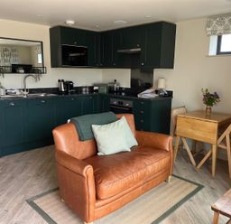 A kitchen with dark green cabinets a small brown leather sofa and a wooden dining table with chairs and flowers at The Cabin at Ropewalk in Goonbell near St Agnes