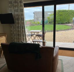 A living room with a leather armchair a wooden table and chairs a wall-mounted television and a sliding glass door opening to a garden at The Cabin at Ropewalk in Goonbell near St Agnes