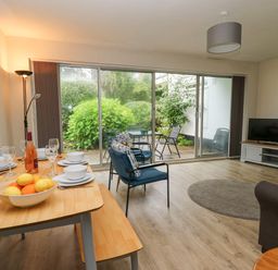 A room with a dining table set with plates glasses and fruit two blue chairs a television on a stand and sliding glass doors opening to a garden at 14 Coedrath Park in Saundersfoot