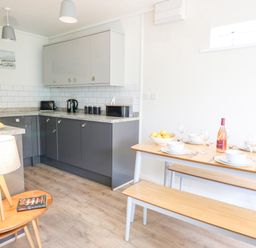 A kitchen with gray cabinets and a wooden dining table with benches set with dishes and a bottle at 14 Coedrath Park Saundersfoot