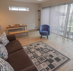 A living area with a brown sofa with patterned cushions a blue armchair a wooden dining table with benches and a floor lamp at 19 Coedrath Park in Saundersfoot