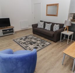 A living room with a gray sofa and patterned cushions a blue armchair a patterned rug a wooden tv stand with a television a side table with a lamp and a wooden dining table at 19 Coedrath Park in Saundersfoot