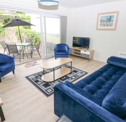 A living room with blue sofa and chairs a coffee table and television with sliding glass doors to a patio at 29 Coedrath Park Saundersfoot