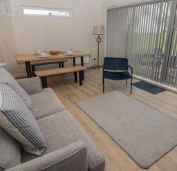 A living room with a gray sofa, blue armchair, wooden dining table with benches, floor lamp, and vertical blinds at 23 Coedrath Park Saundersfoot