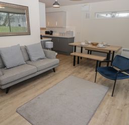 A living area with a gray sofa, a blue armchair, a wooden dining table with bench seats, and a kitchen in the background at 23 Coedrath Park Saundersfoot