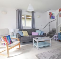 A living room with a gray sofa blue armchair wooden chairs white coffee table and staircase at Rainbow Lodge in St Tudy