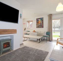 A living room with a wall mounted TV above a fireplace and a dining table with chairs and a bench at Rainbow Lodge in St Tudy
