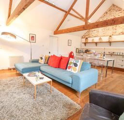 An open plan living room and kitchen with blue sofa wooden beams and a dining table at Packway Barn in Halesworth