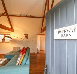 An open door with a sign reading Packway Barn overlooking a living room with a blue sofa and wooden beams at Packway Barn in Halesworth