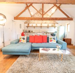 An open living area with a blue sectional sofa red and patterned pillows a white coffee table rug wooden floor and kitchen in the background at Packway Barn in Halesworth