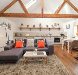 A living room with a gray sectional sofa orange pillows a coffee table and a kitchen with white cabinets and wooden beams at The Nook in Chediston near Halesworth