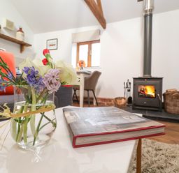 A glass vase with flowers and a book on a table in a living room with a wood stove and baskets at The Nook in Chediston near Halesworth