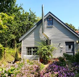 A small house with gray siding surrounded by trees and purple flowering plants at Twosome in Chediston near Halesworth