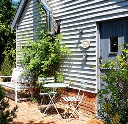 A white bench and two chairs with a round table outside a building with blue door and white siding at Twosome in Chediston near Halesworth