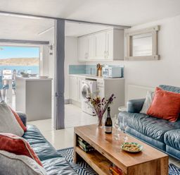 A living room with blue leather sofas, a wooden coffee table, and a kitchen area with white cabinets and a washing machine at The Cubby Hole in Sennen Cove