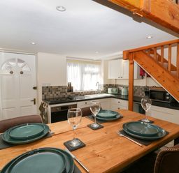 A dining area with a wooden table set with teal plates and wine glasses next to a kitchen with white cabinets and a wooden staircase at Harvest Moon Cottage in Much Cowarne