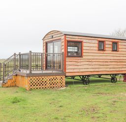 A wooden shepherd hut on wheels with a small deck and railing in a grassy field at Tal Sarn Shepherd Hut in Dwyran near Niwbwrch Newborough