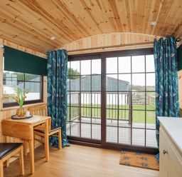 A wooden room with a small table and two chairs near large glass sliding doors with blue patterned curtains at Tal Sarn Shepherd Hut in Dwyran near Niwbwrch Newborough