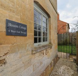 The exterior wall of a stone building with a window a sign saying Blossom Cottage and a metal garden gate at Blossom Cottage in Chipping Campden