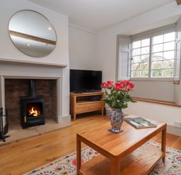 A living room with a fireplace burning fire a round mirror above a mantel a wooden coffee table with a vase of flowers a window with shutters and a television on a wooden stand at Blossom Cottage in Chipping Campden
