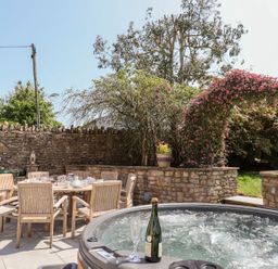An outdoor patio with a wooden dining table and chairs set with dishes and a hot tub with a bottle and glass against a stone wall with plants at Nempnett Farmhouse in Nempnett Thrubwell near Blagdon