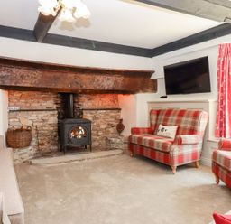 A living room with a stone fireplace and wood stove with plaid armchairs and a wall-mounted television at Nempnett Farmhouse in Nempnett Thrubwell near Blagdon