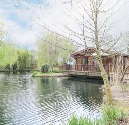 A wooden cabin on stilts beside a pond with trees and greenery at Kingfisher Lodge in Badwell Ash