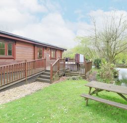 A wooden cabin with a ramp and stairs next to a pond with trees and a picnic table on grass at Kingfisher Lodge in Badwell Ash