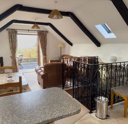 An attic living and dining area with exposed beams a dining table chairs a brown leather sofa and armchair a television telescope and balcony doors at Crogal Stable in New Quay