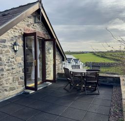 A stone building with open glass doors leading to a roof terrace with a wooden table and chairs overlooking green fields at Crogal Stable in New Quay