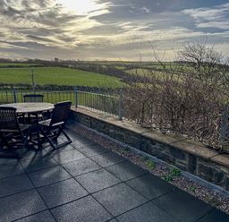 An outdoor patio with a round wooden table and four wooden chairs overlooking green fields and a cloudy sky at Crogal Stable in New Quay