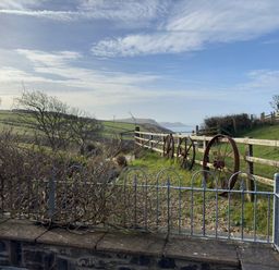 A garden area with a metal fence and wooden fence with wheels alongside a grassy hill overlooking distant hills and sea at Crogal Stable in New Quay
