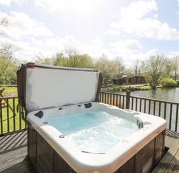 An outdoor hot tub on a wooden deck overlooking a pond with trees and a small cabin in the background at Wagtail Lodge in Badwell Ash