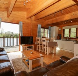 A wooden interior with a seating area with leather sofas and a coffee table a dining area with a table and chairs and a kitchen with white cabinets and windows at Wagtail Lodge in Badwell Ash