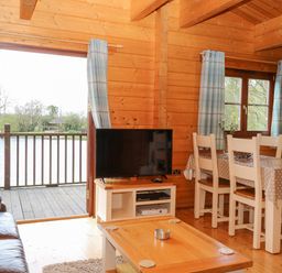 A wooden living room with a leather sofa a television on a stand a dining table with chairs and an open door to a balcony at Wagtail Lodge in Badwell Ash